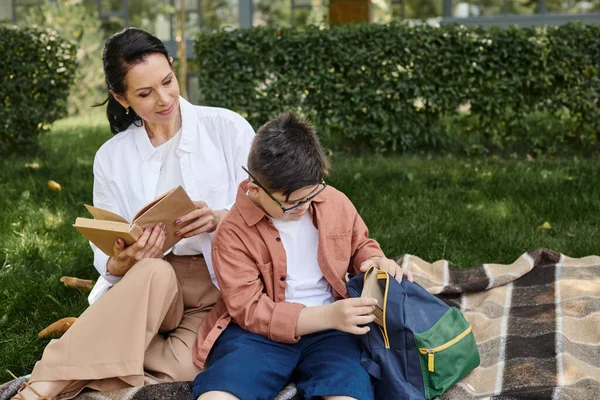 Boy with down syndrome taking off book from school backpack near happy mother on blanket in park — Stock Photo