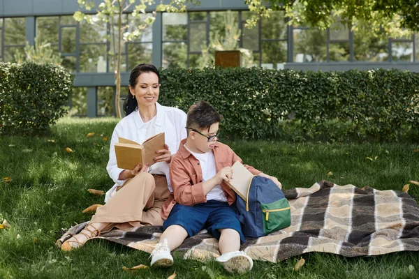 Lächelnde Frau mittleren Alters und ihr Sohn mit Down-Syndrom sitzen mit Buch und Rucksack im Park — Stockfoto