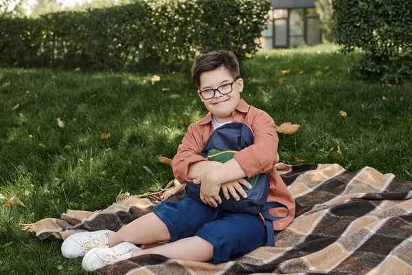 Menino feliz e elegante com síndrome de down sentado perto da mochila da escola em cobertor no parque — Fotografia de Stock