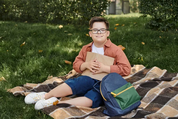 Glücklicher Junge mit Down-Syndrom hält Buch in der Nähe von Schulrucksack auf Decke im Park — Stockfoto