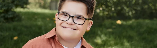 Cheerful and genuine kid with down syndrome in eyeglasses smiling in park, portrait, banner — Stock Photo