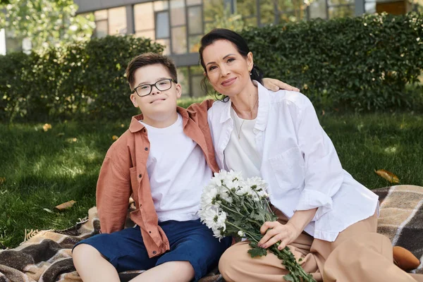 Frau mittleren Alters hält Blumen in der Nähe ihres Sohnes mit Down-Syndrom auf Decke im Park — Stockfoto