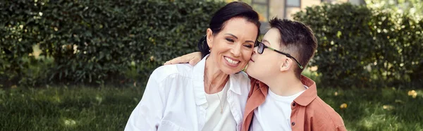 Preteen boy with down syndrome kissing cheerful mother in park, emotional connection, love, banner — Stock Photo