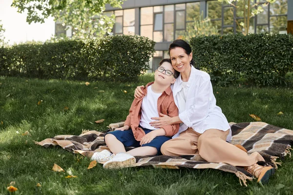 Усміхнена жінка середнього віку, що приймає дев'ятнадцятирічного сина з синдромом Дауна на ковдрі в парку, якісний час — стокове фото