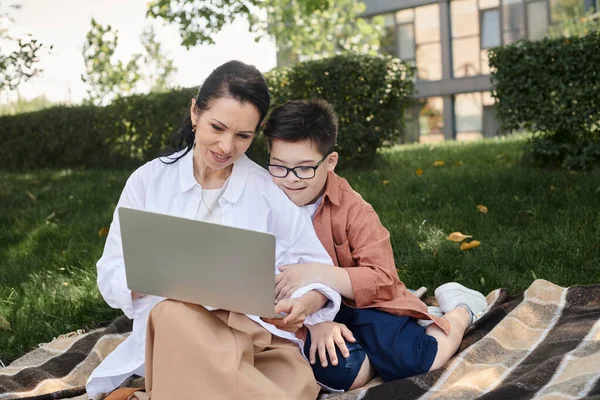 Happy middle aged woman sitting with laptop near son with down syndrome on blanket in park — Stock Photo