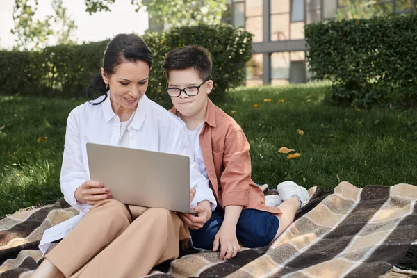 Ребенок с синдромом Дауна смотрит на ноутбук рядом с улыбающейся матерью на одеяле в парке, электронное обучение — стоковое фото