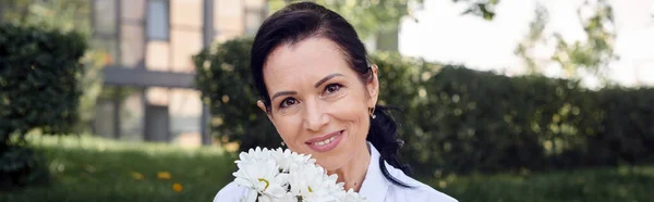 Despreocupada mujer de mediana edad sosteniendo flores y sonriendo a la cámara en el parque, retrato, pancarta - foto de stock