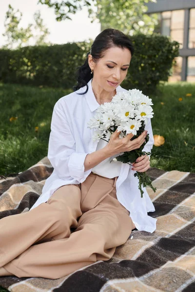 Heureux femme d'âge moyen en tenue décontractée élégant tenant des fleurs tout en se reposant sur la couverture dans le parc — Photo de stock