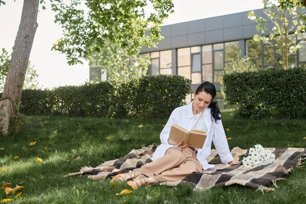 Жінка середнього віку в повсякденному вбранні читає роман, сидячи на ковдрі в парку, якісний час — стокове фото