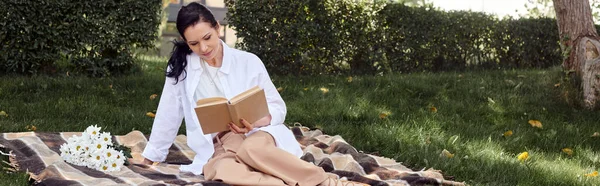 Middle aged woman in casual attire sitting on blanket in park and reading book, banner — Stock Photo