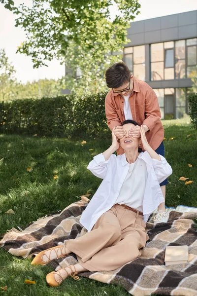 Малыш с синдромом Дауна веселится и играет угадай, кто играет с матерью, сидящей на одеяле в парке — стоковое фото