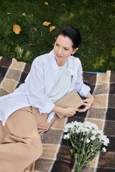 Top view of positive middle aged woman in casual attire looking away near book on blanket in park — Stock Photo
