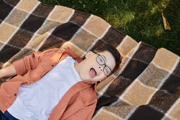 Top view of carefree boy with down syndrome, in eyeglasses, sticking out tongue on blanket in park — Stock Photo