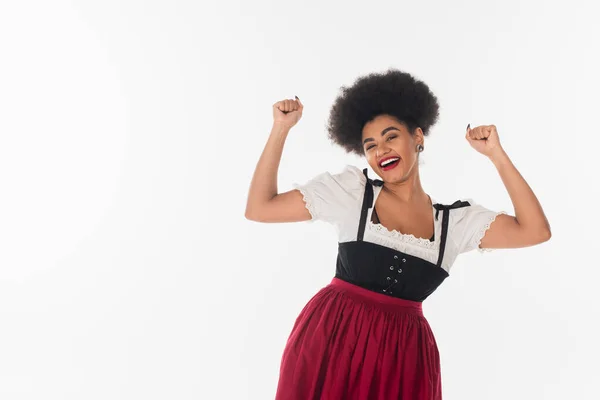 Excited african american waitress in traditional bavarian costume rejoicing and gesturing on white — Stock Photo