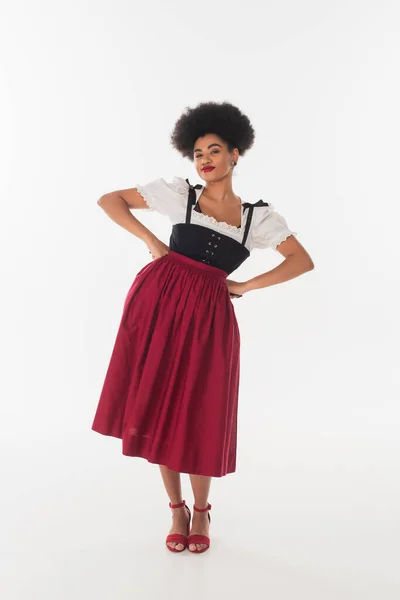 Щаслива афроамериканська баварська офіціантка в автентичному одязі, позує руками на стегнах на білому — стокове фото