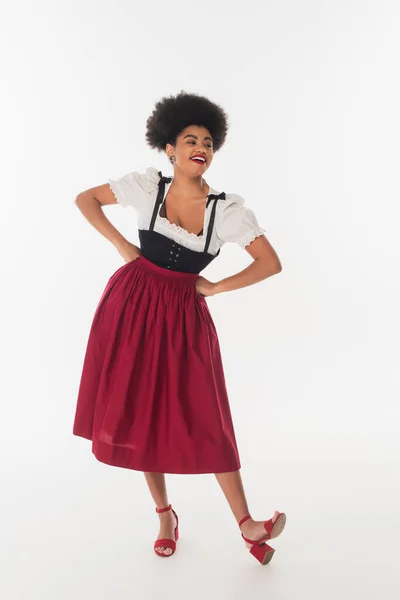 Весела афроамериканська американська офіціантка в дирндлі, дивлячись з руками на стегнах на білому — стокове фото