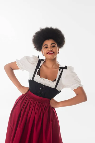 Joyful african american oktoberfest waitress in traditional dirndl dress with hands on hips on white — Stock Photo