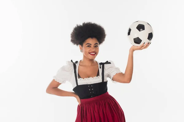 Smiling african american bavarian waitress in dirndl with soccer ball on white, oktoberfest concept — Stock Photo