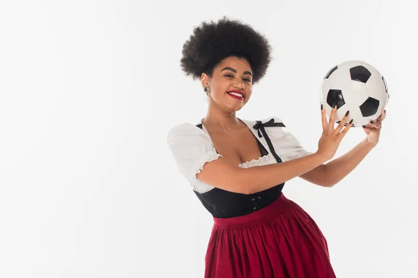 Alegre octoberfest camarera en traje bavariano celebración de pelota de fútbol afroamericano en blanco - foto de stock