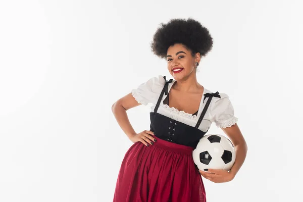 African american oktoberfest waitress in bavarian costume with soccer ball and hand on hip on white — Stock Photo