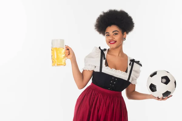 Africano americano oktoberfest camarera con pelota de fútbol y taza de cerveza lager con espuma en blanco - foto de stock