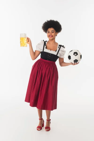 Serveuse bavaroise afro-américaine posant avec ballon de football et tasse de bière artisanale avec mousse sur blanc — Photo de stock
