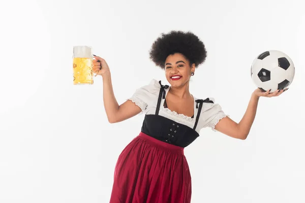 Щаслива афроамериканська офіціантка з чашкою пива з піною та футбольним м'ячем на білому — стокове фото