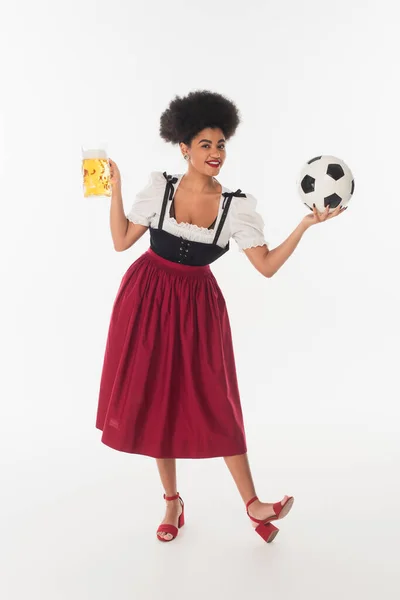African american oktoberfest waitress with mug of foamy beer playing football on white, full length — Stock Photo