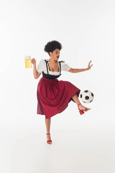 Cheerful african american bavarian waitress with mug of foamy bear stepping on soccer ball on white — Stock Photo