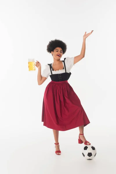 Joyeuse serveuse afro-américaine oktoberfest avec tasse de bière mousseuse artisanale près du ballon de football sur blanc — Photo de stock