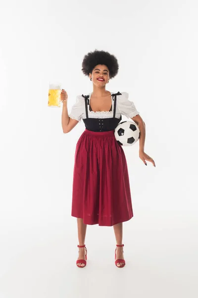 Serveuse bavaroise afro-américaine avec ballon de football et tasse de bière souriante sur blanc, oktoberfest — Photo de stock
