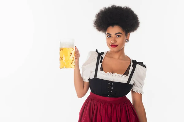 Pleased african american octoberfest waitress in authentic costume with mug craft beer on white — Stock Photo