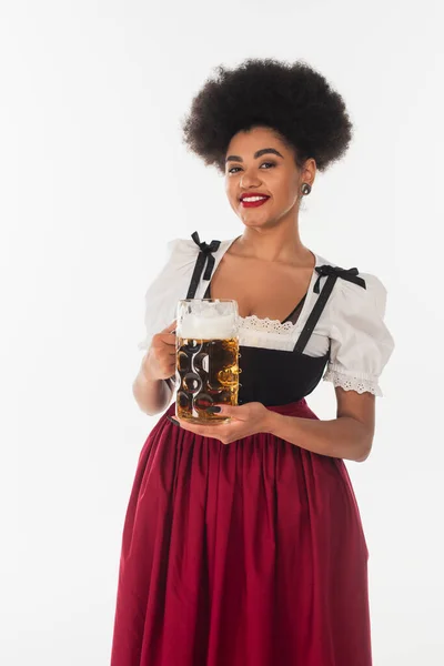 Encantadora camarera bavariana afroamericana en vestido tradicional sosteniendo taza de cerveza espumosa en blanco - foto de stock
