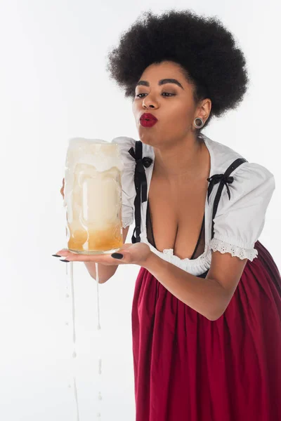 Serveuse bavaroise afro-américaine en dirndl soufflant de la mousse de bière dans une tasse sur blanc, oktoberfest — Photo de stock