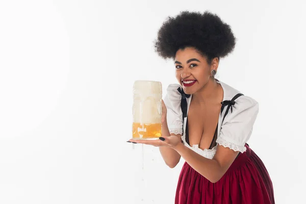 African american oktoberfest waitress holding mug of beer with foam and looking at camera on white — Stock Photo