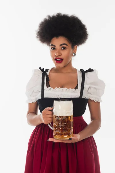 Amazed african american bavarian waitress in oktoberfest costume with mug of foamy beer on white — Stock Photo