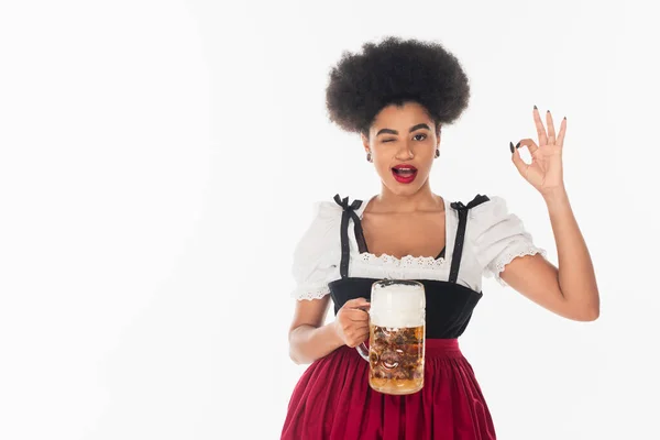 African american oktoberfest waitress with mug of craft beer winking and showing okay sign on white — Stock Photo