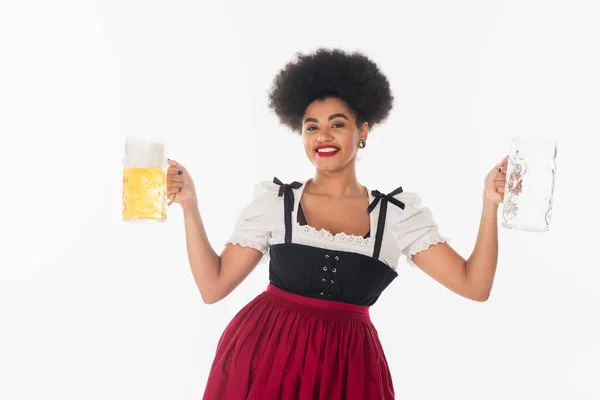 African american bavarian waitress in oktoberfest dirndl with empty and full beer mugs on white — Stock Photo