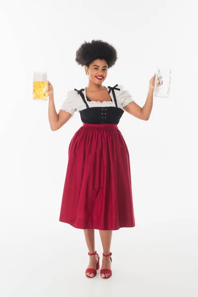 Cameriera afroamericana oktoberfest in costume tradizionale con tazze di birra vuote e piene su bianco — Foto stock
