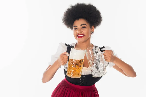 Pleased african american bavarian waitress with full and empty beer mugs on white, oktoberfest — Stock Photo