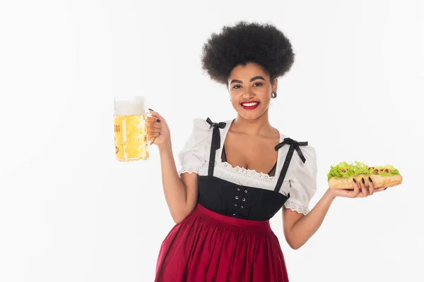 Cheerful african american bavarian waitress with beer mug and hot dog on white, oktoberfest concept — Stock Photo