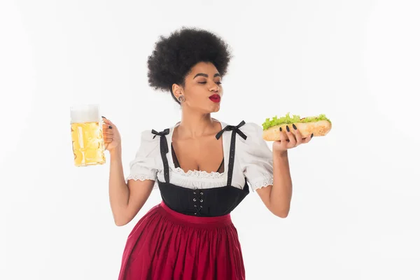 Delighted african american oktoberfest waitress in dirndl with tasty hot dog and beer mug on white — Stock Photo