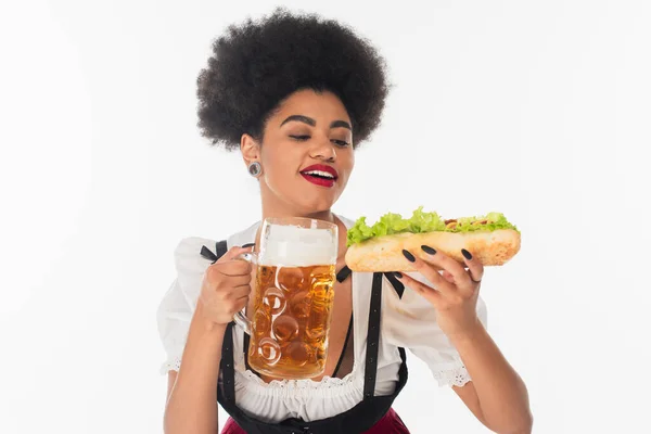 Улыбающаяся африканская американская баварская официантка с кружкой пива и вкусным хот-догом на белом, октоберфест — стоковое фото