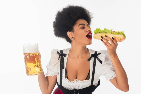 Serveuse bavaroise afro-américaine avec tasse de bière mangeant un hot dog savoureux sur blanc, Oktoberfest — Photo de stock