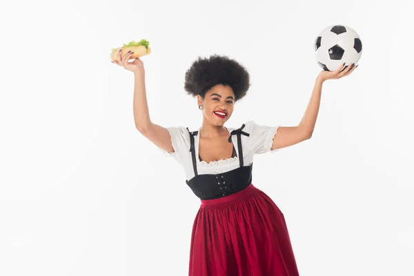 Excitée serveuse bavaroise afro-américaine avec ballon de football et hot dog dans les mains levées sur blanc — Photo de stock