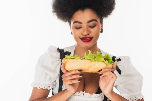 Delighted african american oktoberfest waitress in traditional costume with tasty hot dog on white — Stock Photo