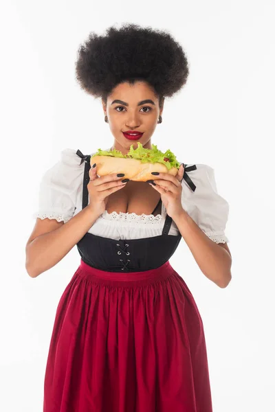 Africano americano oktoberfest camarera en bavarian traje celebración hot dog y sonriendo en blanco - foto de stock