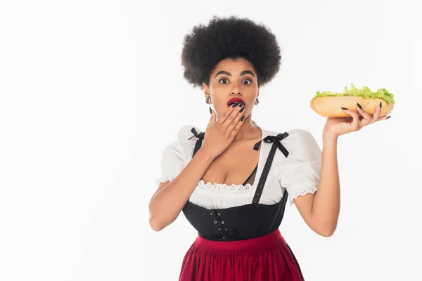 Sorprendida camarera bavariana afroamericana sosteniendo hot dog y cubriendo la boca con la mano en blanco - foto de stock
