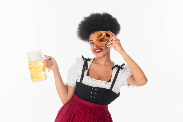 Cheerful african american oktoberfest waitress with mug of beer obscuring face with pretzel on white — Stock Photo