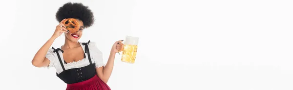 Happy african american bavarian waitress with beer mug obscuring face with pretzel on white, banner — Stock Photo